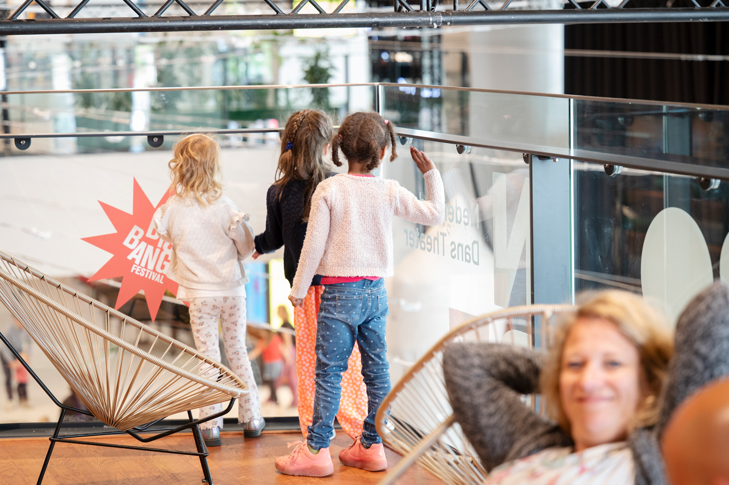 Een reportagefoto van een groepje kinderen die kijken door het glas naar een voorstelling terwijl mama op de voorgrond ontspant. 